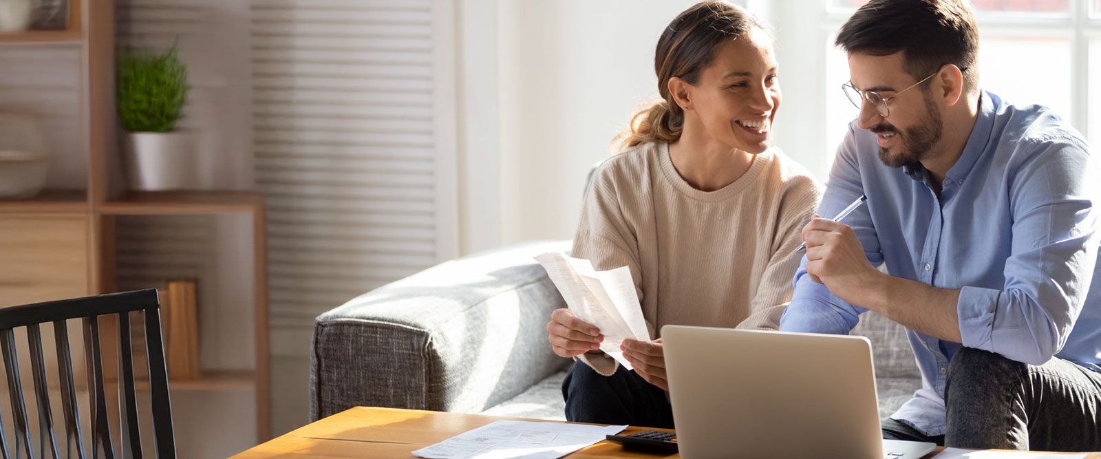 Happy couple sitting on couch reviewing documents while using laptop.