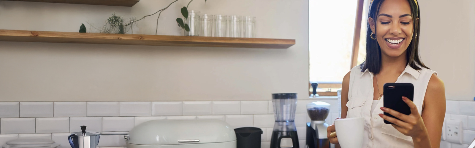 Happy woman standing in her kitchen with a cup of coffee doing her banking on her phone.