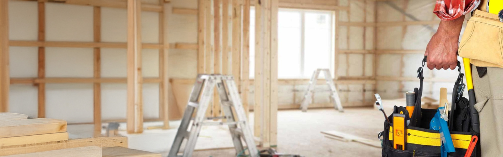 Photo of an empty room with stud framing under construction with a person holding a toolbox in the foreground.